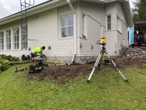 Man utför injektering av geopolymer vid husgrund, en av lösningarna för sättningsproblem. Intill en lasermätare och verktyg.